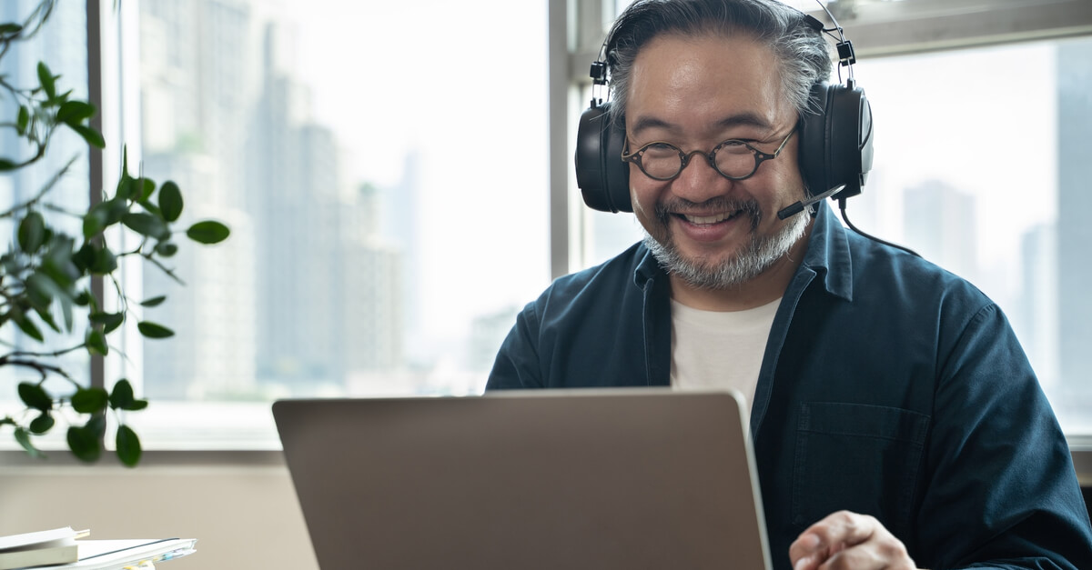 A smiling employee wearing headphones and glasses works at his laptop